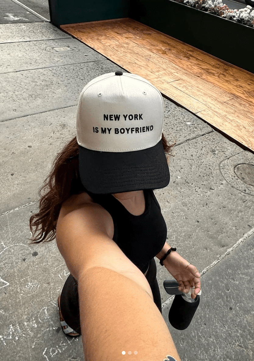 Woman taking a selfie wearing a white and black cap that says "New York Is My Boyfriend."