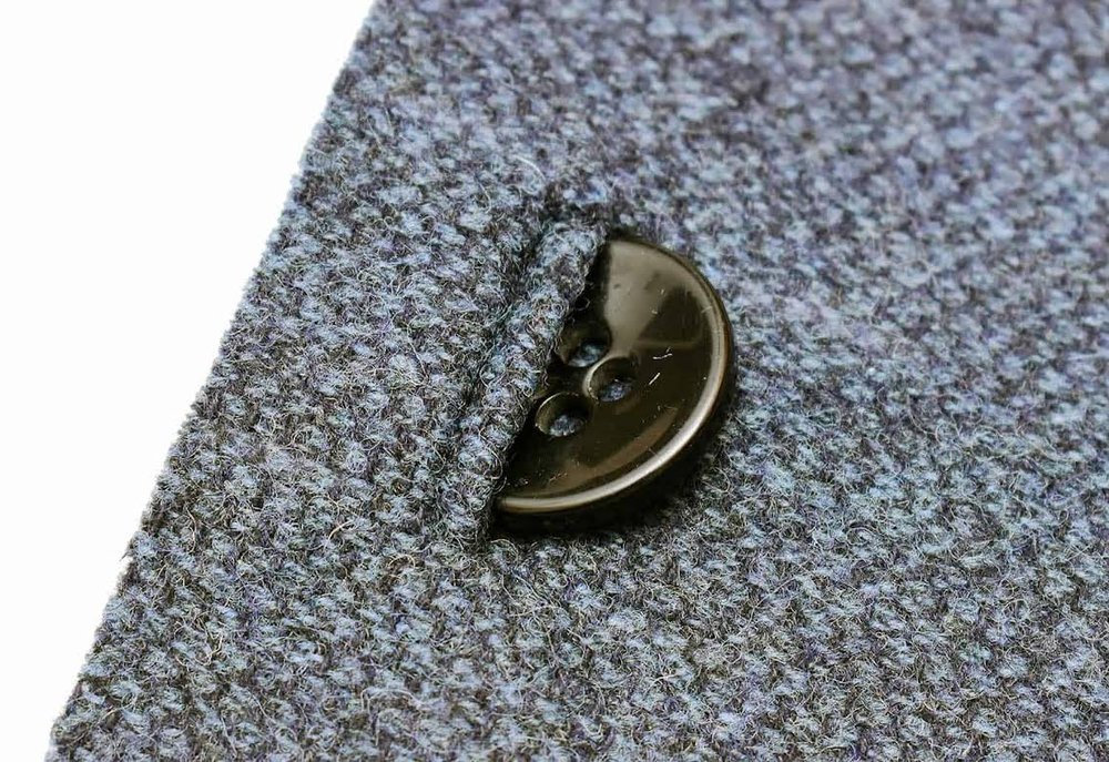 Close-up of a dark button in a horizontal buttonhole on gray houndstooth fabric.