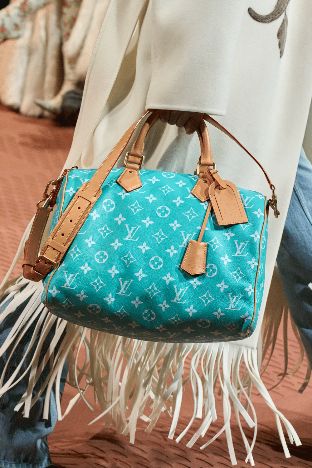 Close-up of a turquoise Louis Vuitton monogram handbag with tan leather straps and gold hardware, worn over a fringed white garment.
