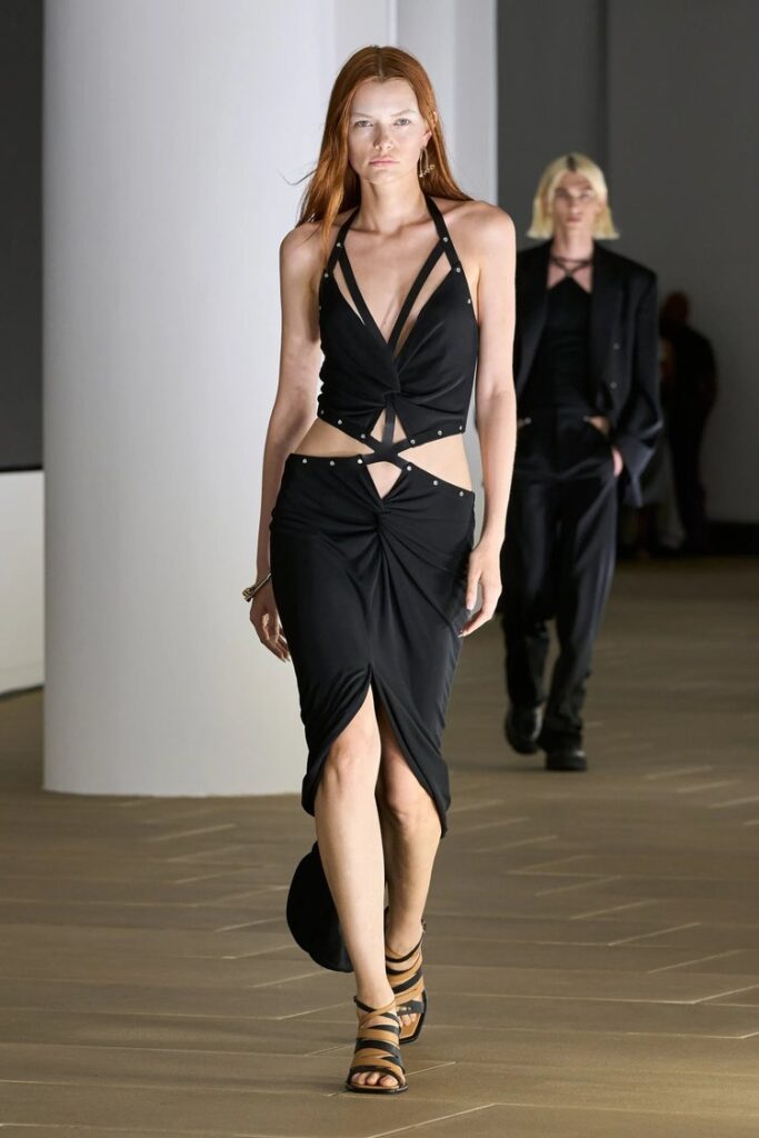 Model walking runway in a cut-out black dress with strappy sandals.