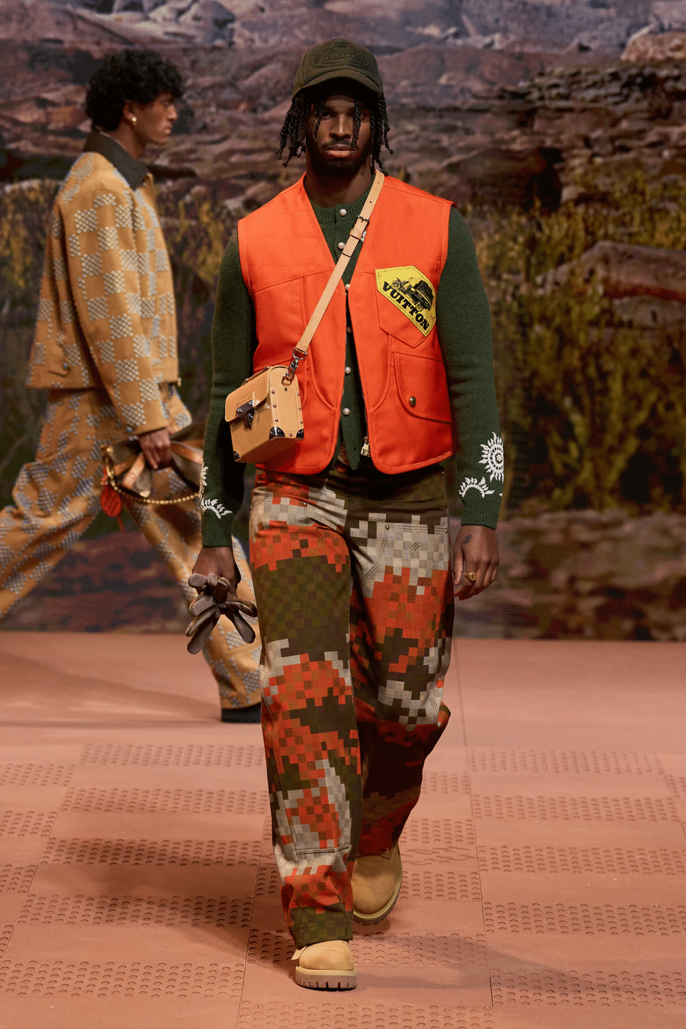 Male model on the runway wearing pixelated camouflage pants, an orange utility vest, green top, and crossbody bag from Louis Vuitton.