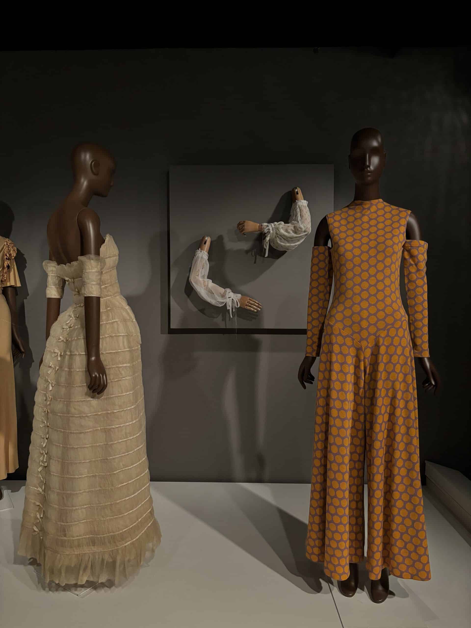 Two mannequins display Stephen Burrows designs—a cream pleated off-shoulder gown and an orange patterned long-sleeve jumpsuit—with sculptural white sleeves framed on the wall behind them.