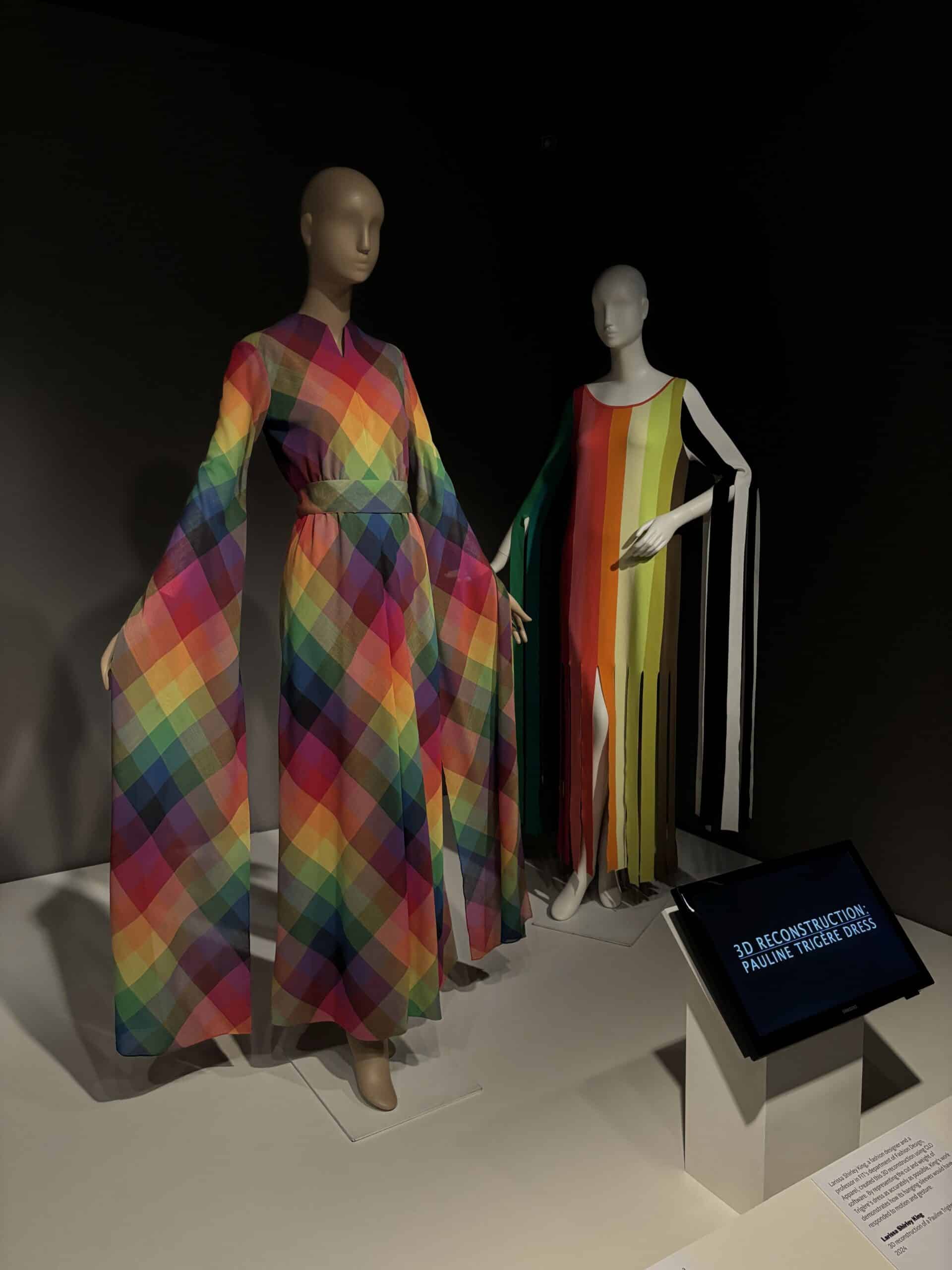 Two multicolor dresses on mannequins, one in a vibrant rainbow check pattern and the other in vertical rainbow stripes with flowing ribbon-like sleeves.