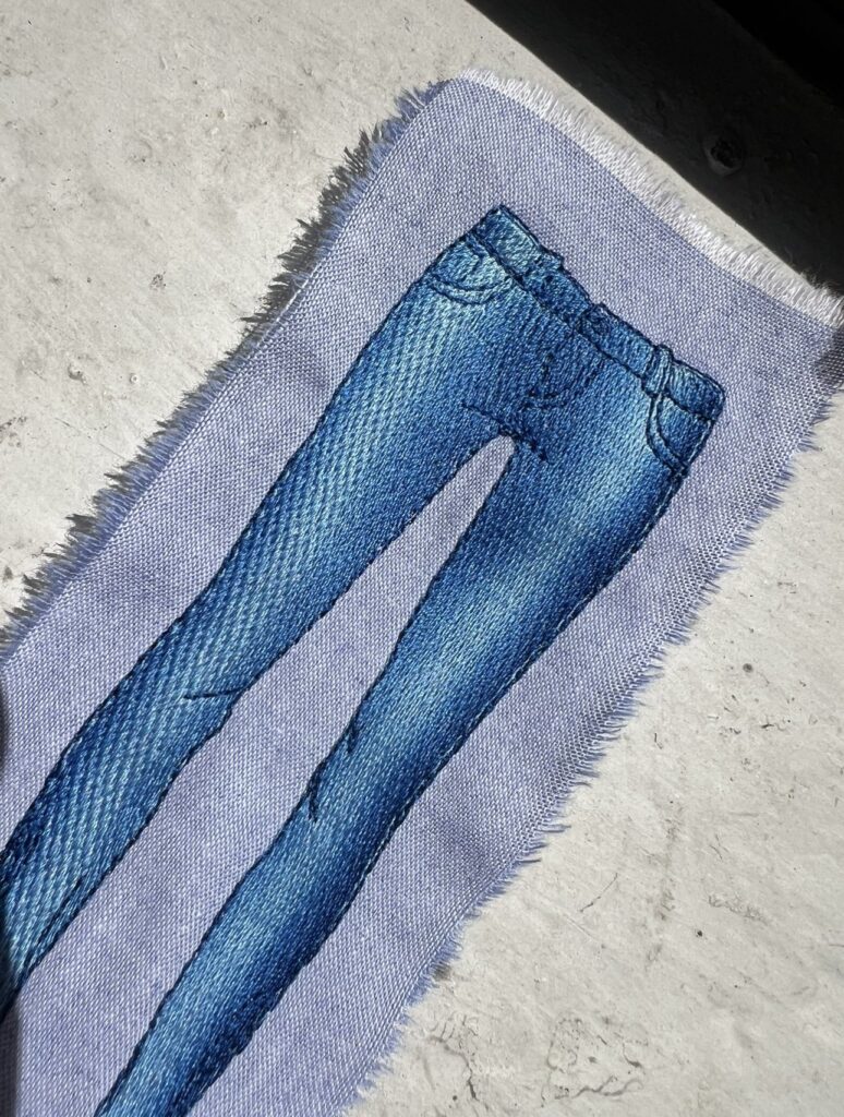 Embroidered design of blue jeans stitched onto light denim fabric, with visible thread texture and shading.