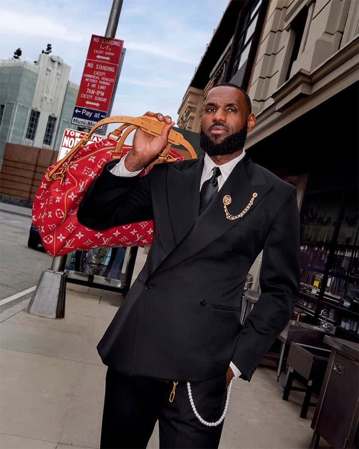 LeBron James walks on a city sidewalk wearing a black double-breasted suit with gold and silver chains, holding a large red Louis Vuitton monogram duffle bag over his shoulder.