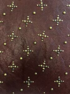 Close-up of maroon leather embellished with small metallic studs arranged in a decorative cross pattern. Detailed image of dark maroon leather featuring small gold and brass-toned studs arranged in cross motifs, creating a textured and ornate surface.