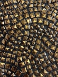 Detailed image of a textile covered with gold-toned pyramid studs in various sizes, arranged in dense swirling clusters on a dark fabric background. Close-up of metallic gold pyramid studs arranged in dense patterns on a dark textile base.