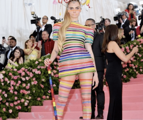 Cara Delevingne wearing a colorful, striped bodysuit and headpiece at the Met Gala, holding a rainbow cane on the pink carpet.