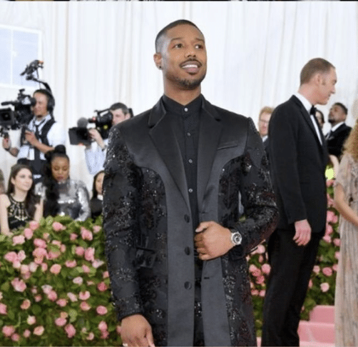 Michael B. Jordan wearing an all-black suit with sequin embellishments and a long overcoat on the Met Gala pink carpet.