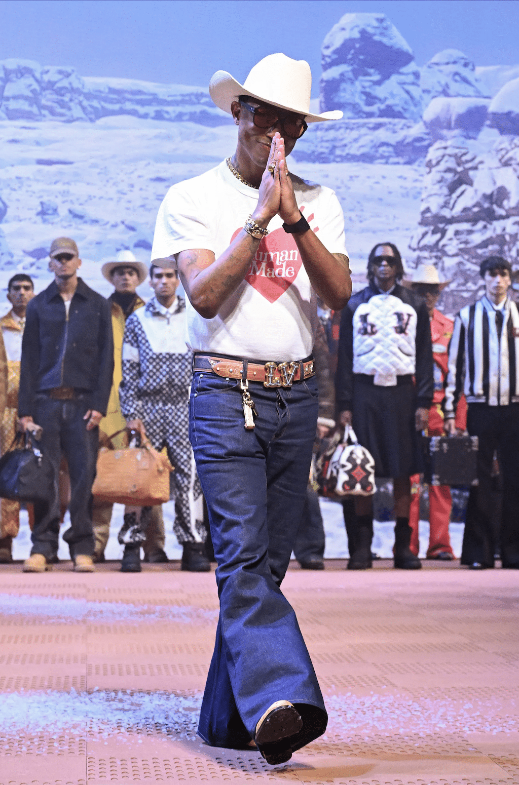 Pharrell Williams walks the runway in a white cowboy hat, graphic t-shirt, flared jeans, and boots, with models standing behind him in Louis Vuitton outfits.