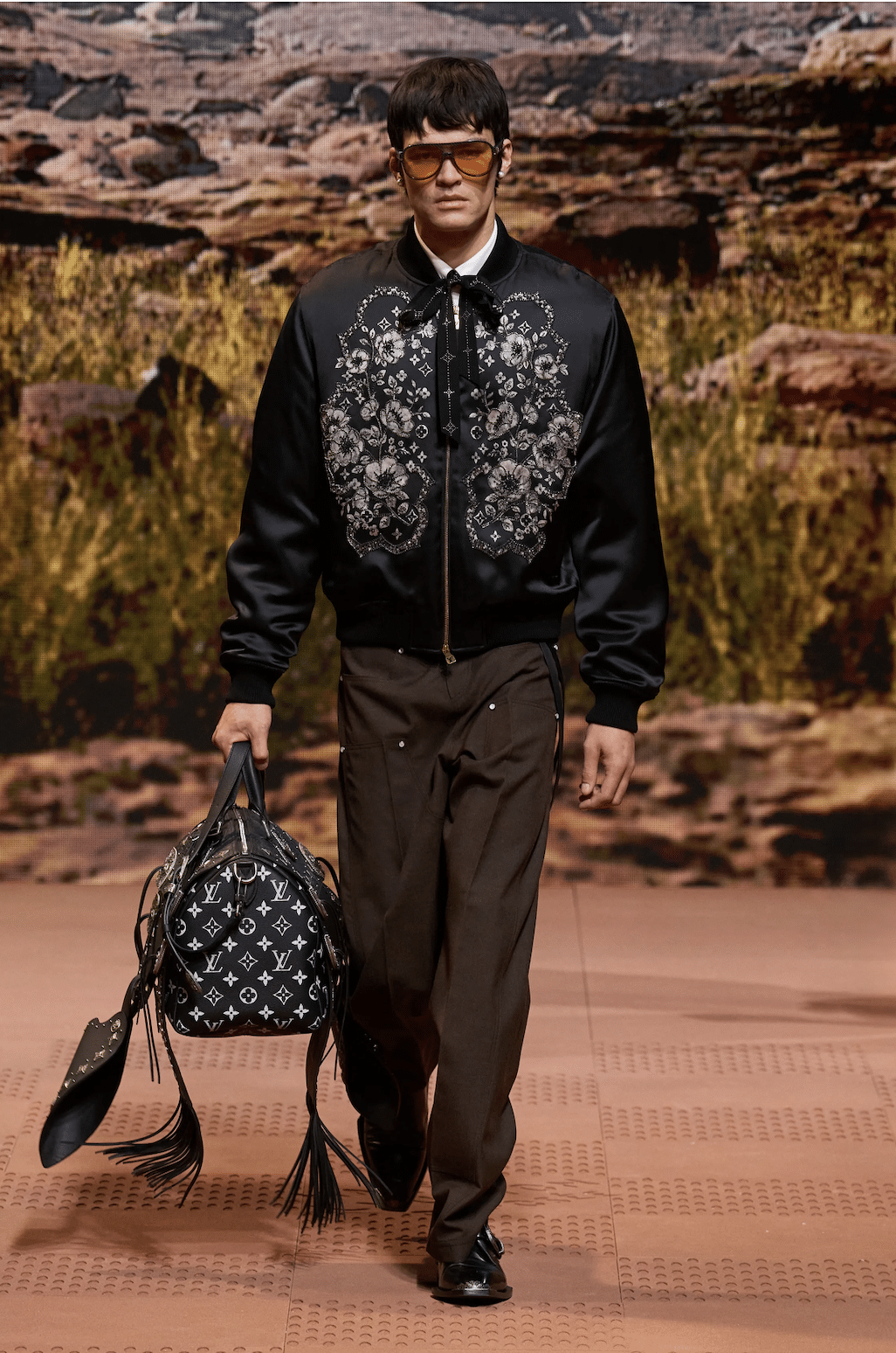Male model wearing a black embroidered bomber jacket with brown pants, carrying a Louis Vuitton monogram bag on the runway.