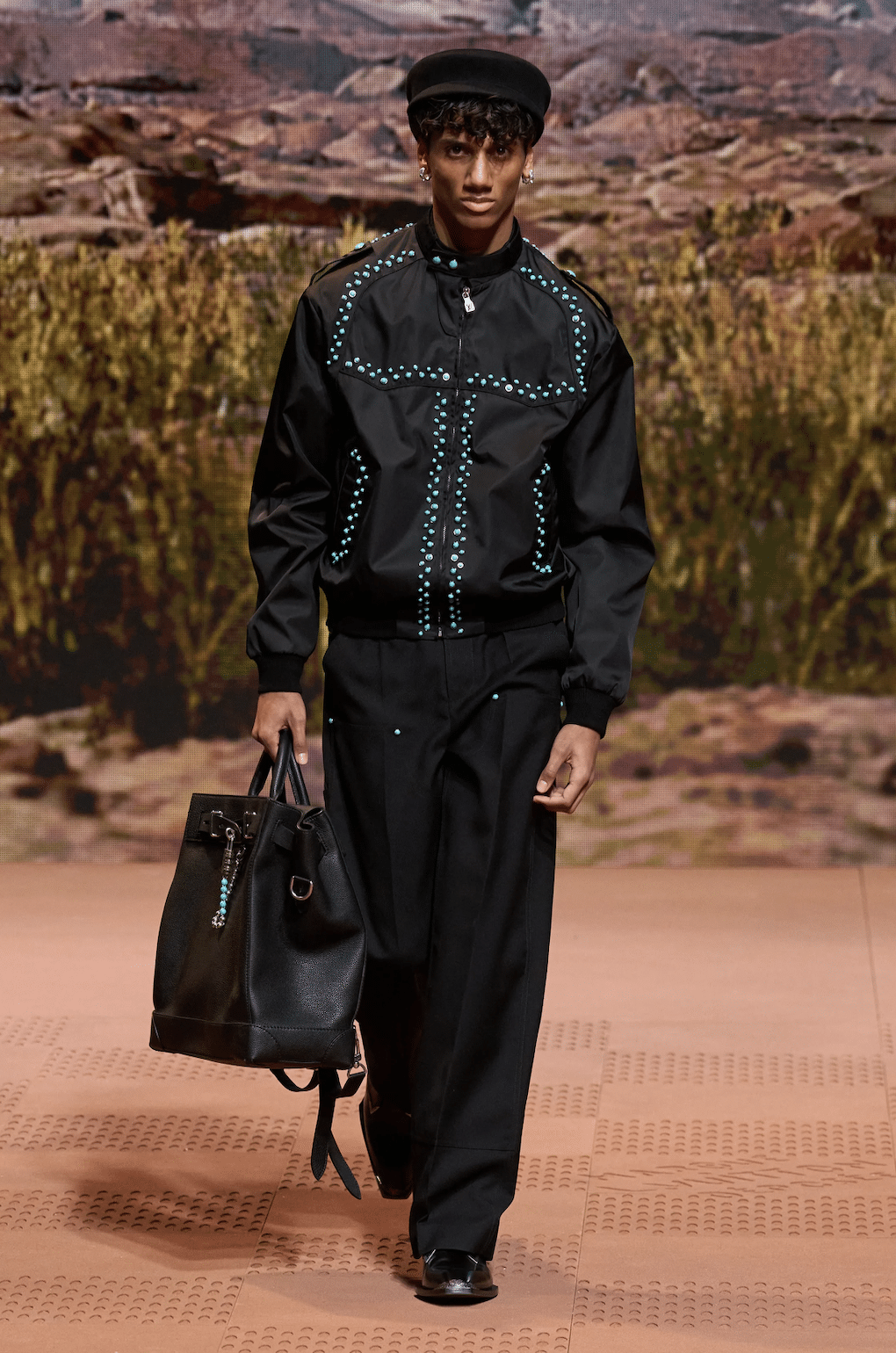 Male model wearing a black Louis Vuitton outfit with turquoise-stitched western detailing, wide-leg pants, and carrying a large black leather bag.