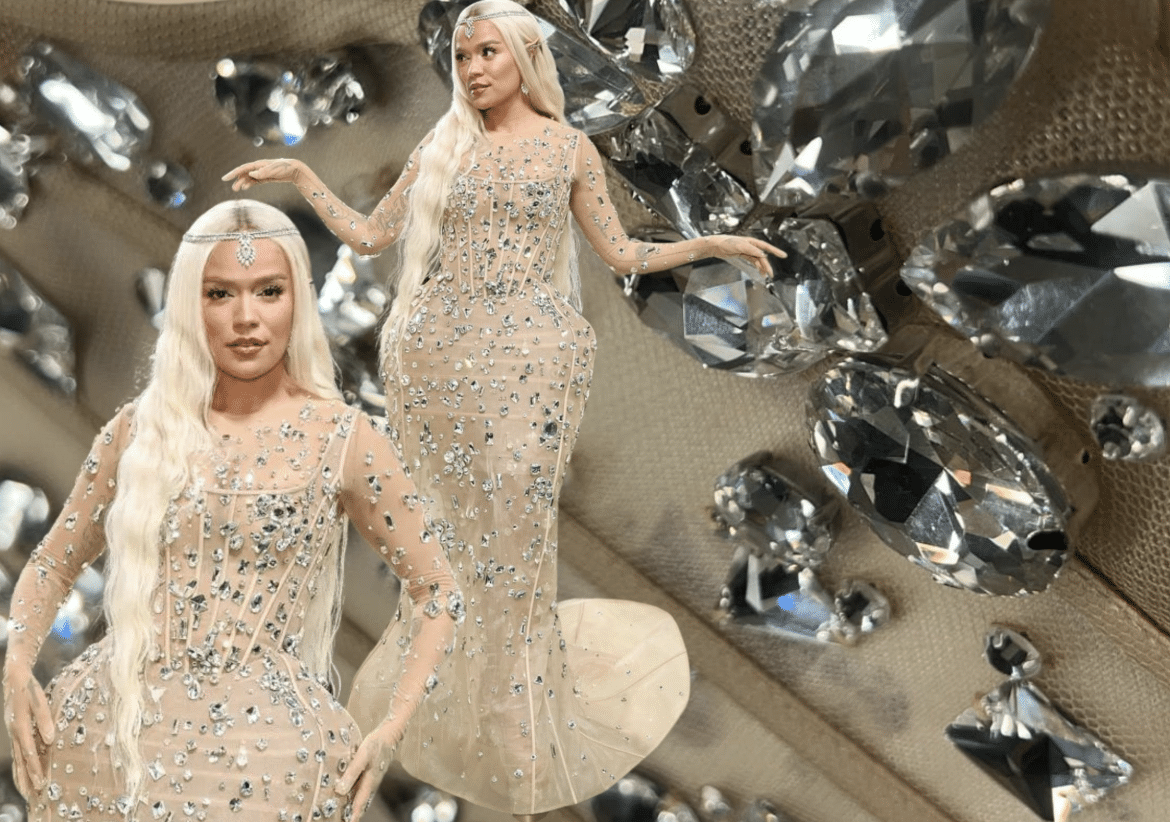 Karol G wearing a sheer nude gown adorned with scattered crystals, set against a close-up background of oversized rhinestones.
