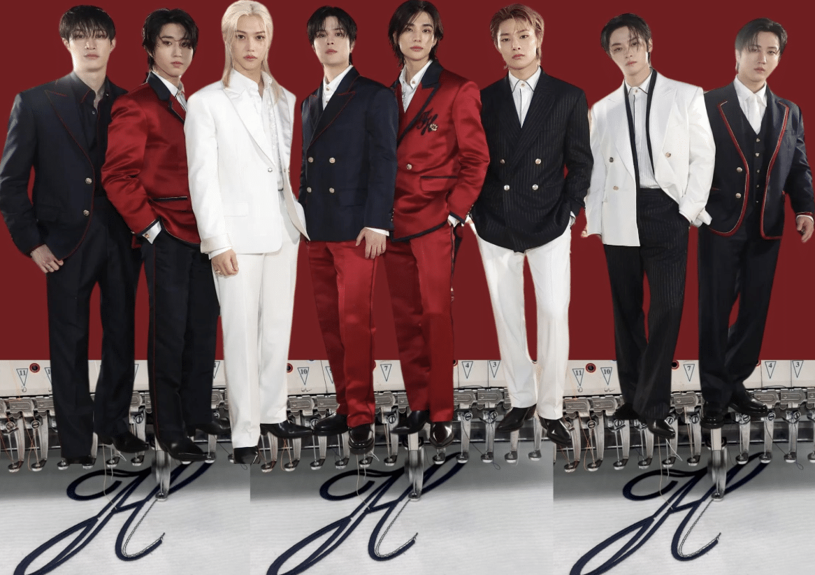Seven members of a K-pop group posing in coordinated red, white, and navy suits against a red backdrop with reflective flooring.