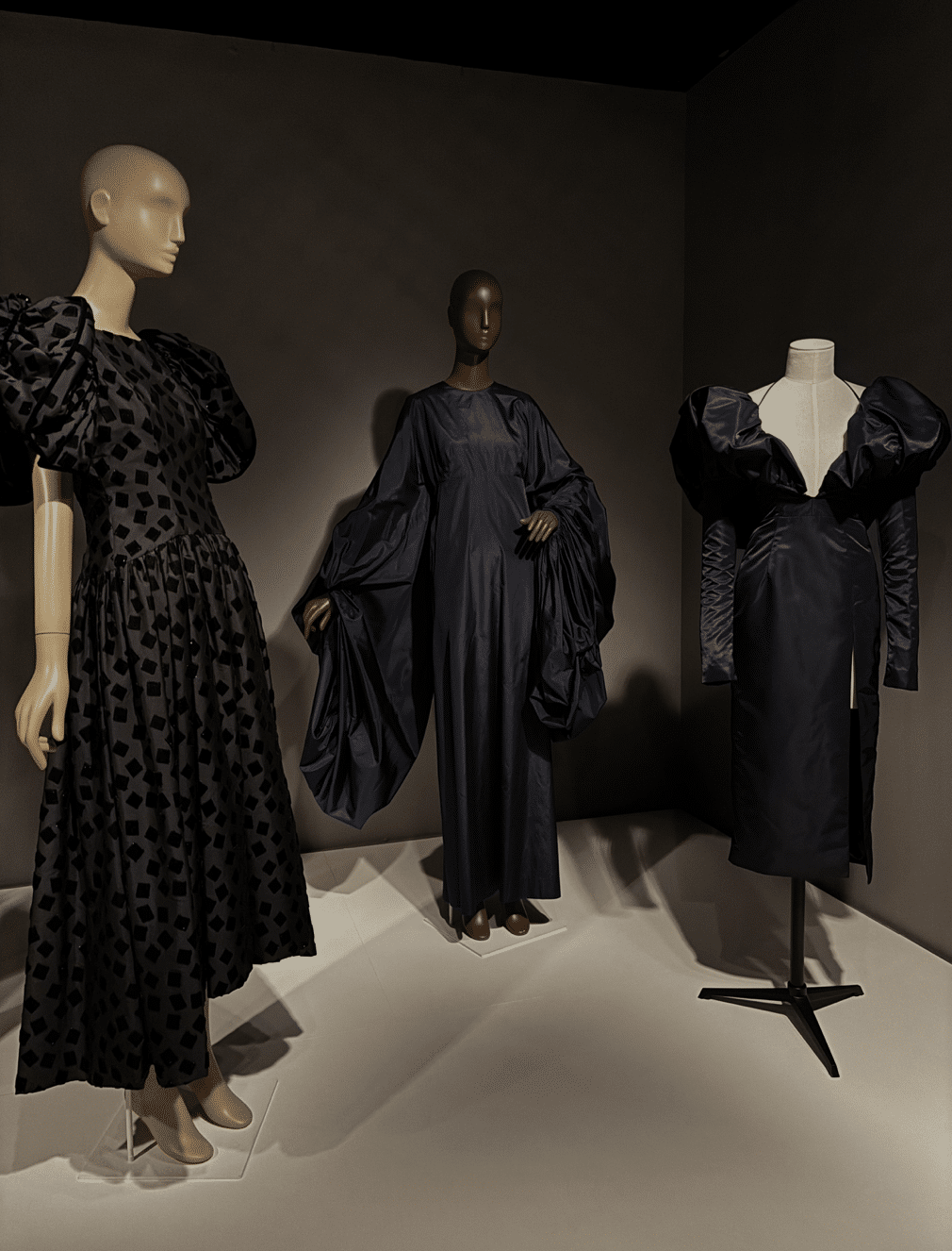 Three black evening dresses displayed on mannequins: one in black silk cut velvet, one in flowing navy silk taffeta, and one in navy acetate faille with exaggerated shoulders.