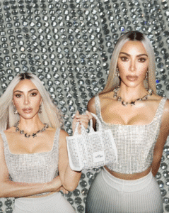 Kim Kardashian wearing a crystal-studded silver tank top and a diamond necklace, holding a sparkling white mini bag in a glitzy backdrop of rhinestones.
