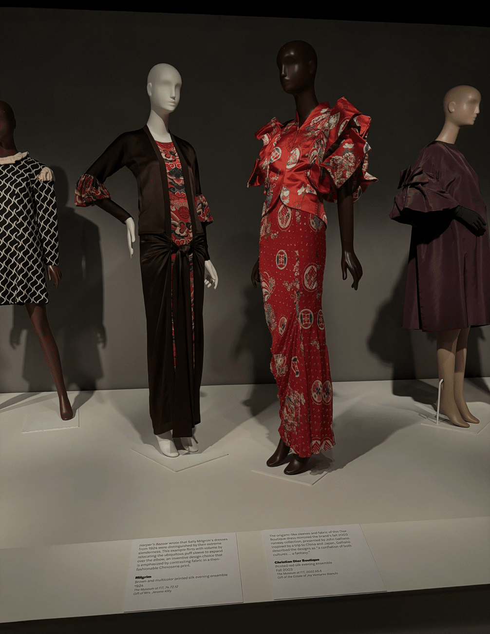Four mannequins display dramatic evening ensembles, including printed silks, bold sleeves, and ruffled details in deep purple, red, black, and multicolor.