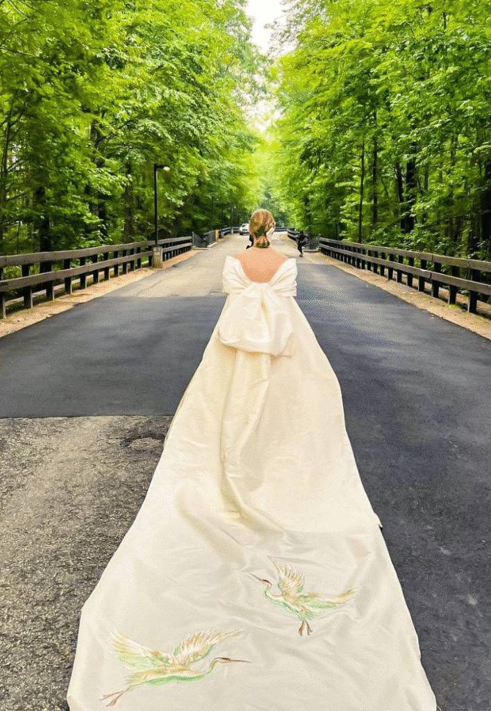 A bride in an elegant ivory gown with a large bow and long train walks down an empty forest road.
