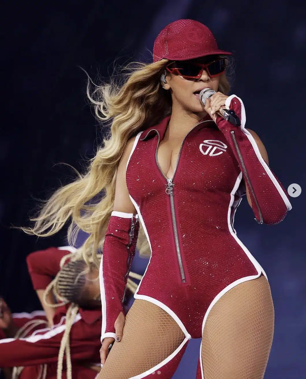 Beyoncé wearing a sparkling maroon Telfar bodysuit with matching hat, visor sunglasses, and arm sleeves while singing on stage.