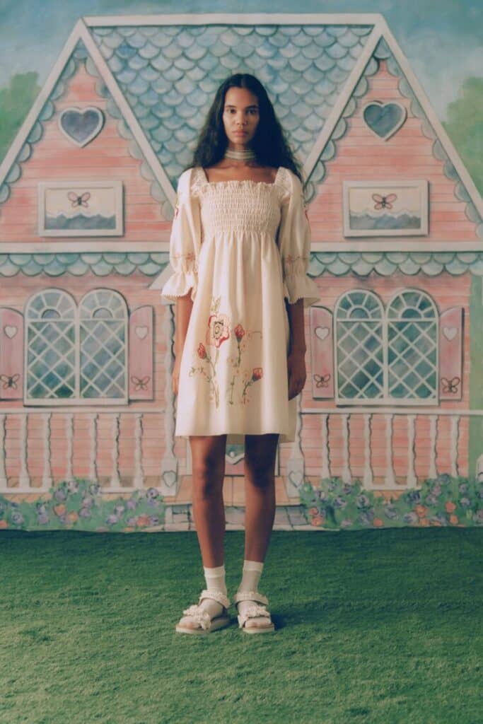 Model wearing a light cream smocked dress with floral embroidery, standing in front of a pink cottage backdrop.