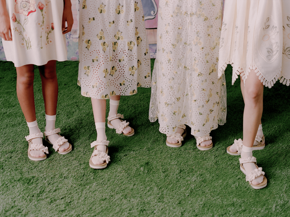 Close-up of four models wearing white dresses and frilly sandals with socks on green grass.