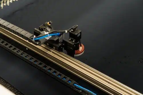 Close-up of a laser cutting head moving across dark fabric on a CNC machine at NYES