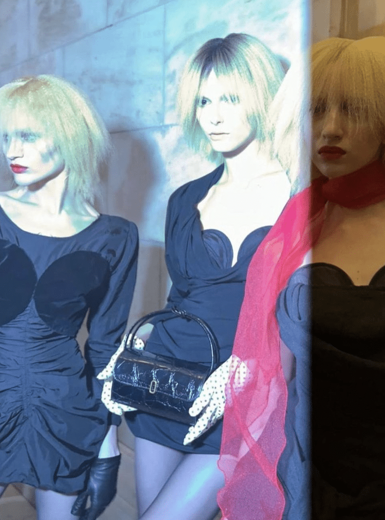Three women in edgy black outfits with dramatic lighting, styled with platinum blonde wigs, red lips, and a vintage black handbag.