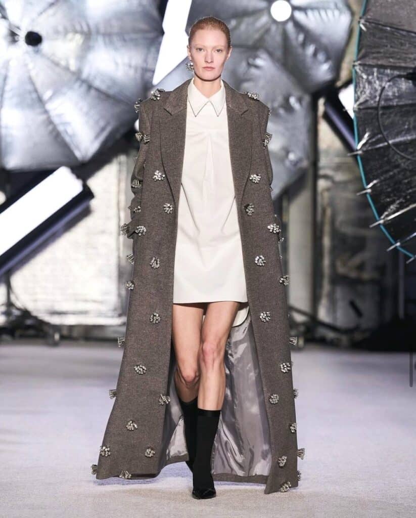 Model in a long embellished coat over a white mini dress, walking a fashion runway.