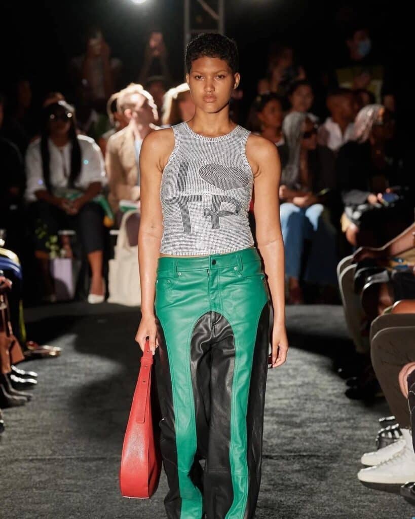 Model wearing a rhinestone-covered tank top reading “I ❤️ TP” paired with two-tone green and black leather pants and holding a red bag on the runway.