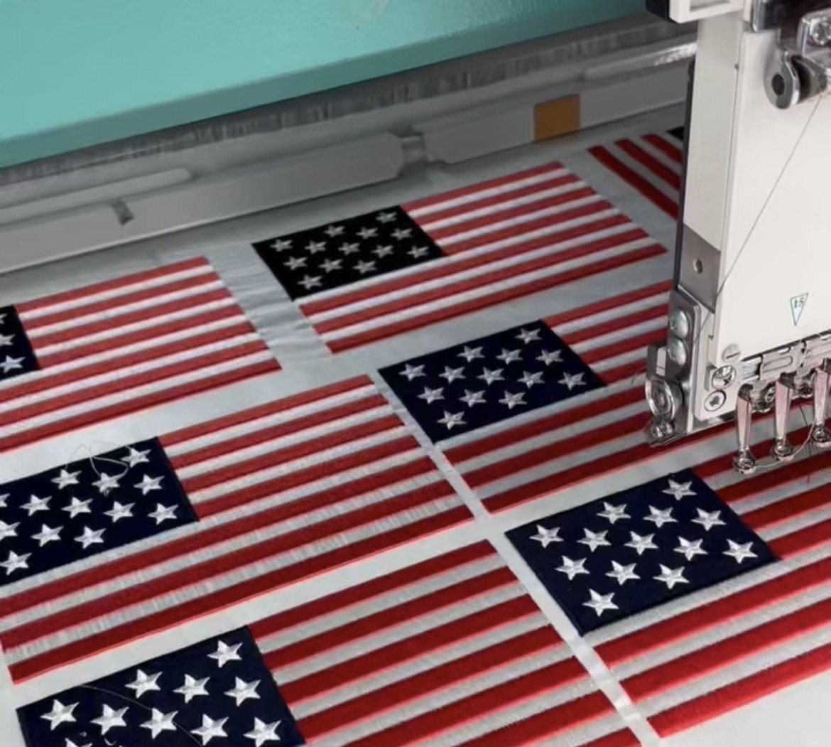 Embroidery machine stitching multiple American flag patches on white fabric.