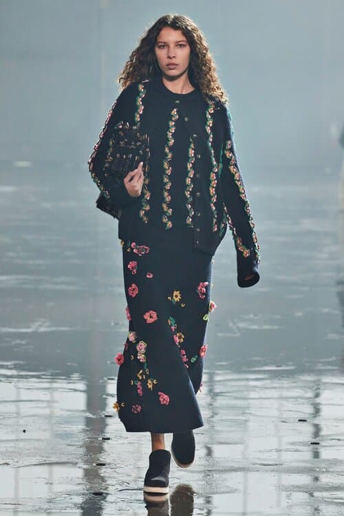 Model in black knit sweater and skirt decorated with colorful floral embroidery.