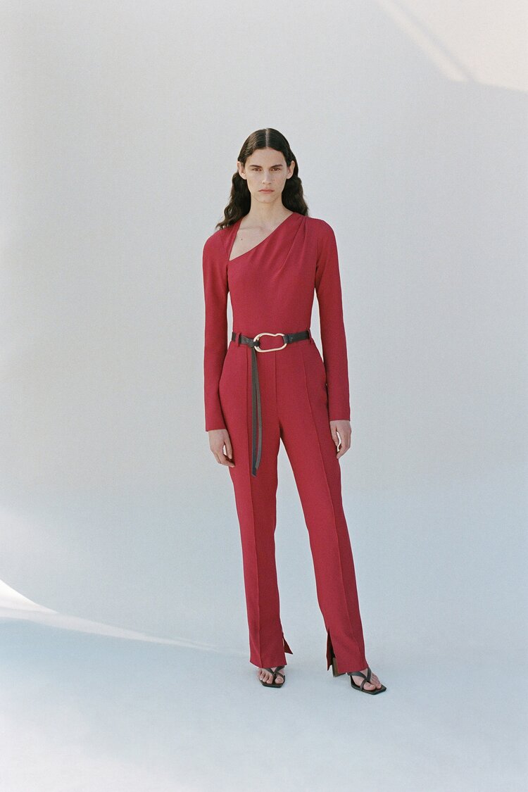 Model wearing a red asymmetrical jumpsuit with long sleeves, cinched at the waist with a black belt, standing against a minimal light background.